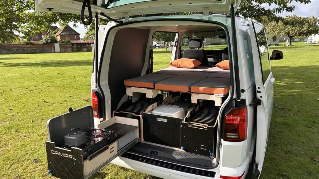 Campal Elevate removable camper conversion kit installed in a white VW Transporter T6.1, featuring a slide-out kitchen with gas stove and a deployed double bed with orange cushions.