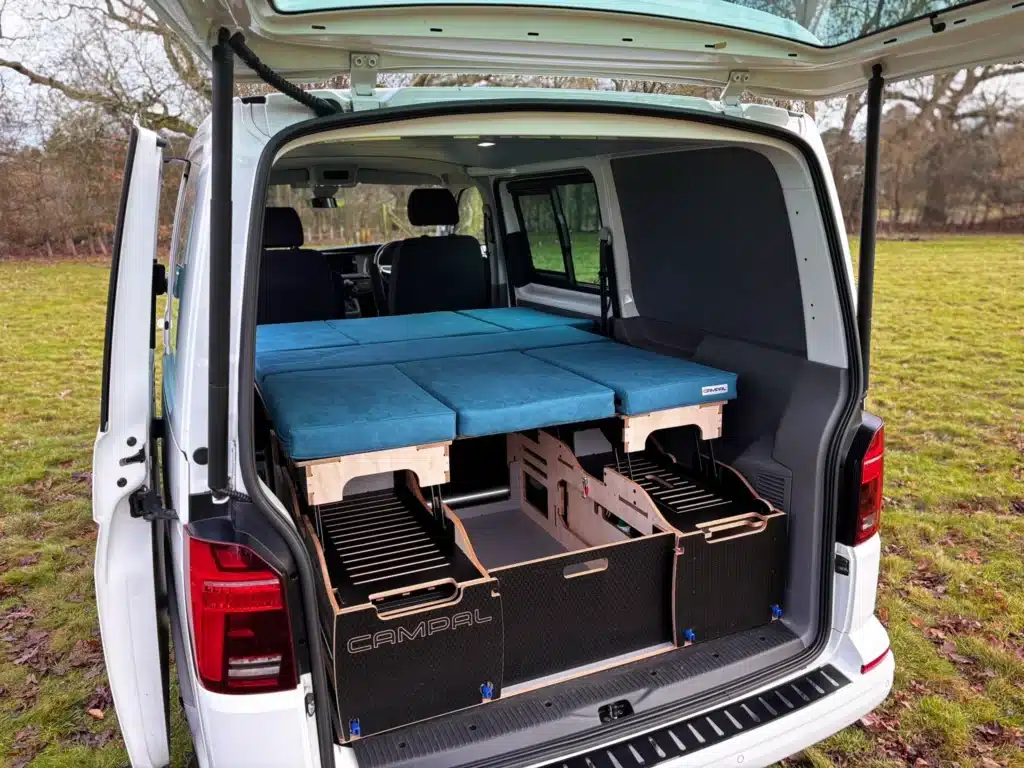 Rear view of a VW Transporter with tailgate open, displaying the Campal Elevate removable camper unit with blue bed cushions and birch plywood storage compartments.