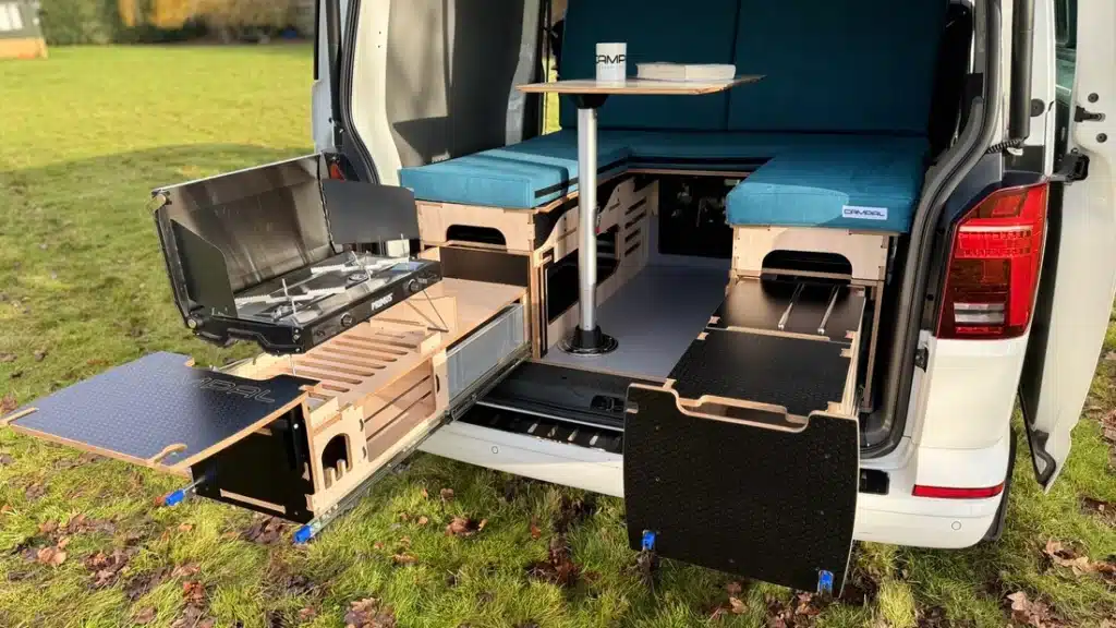 Campal Elevate camper system in a VW Transporter featuring a pull-out outdoor kitchen drawer with Primus stove and an interior dining table setup.