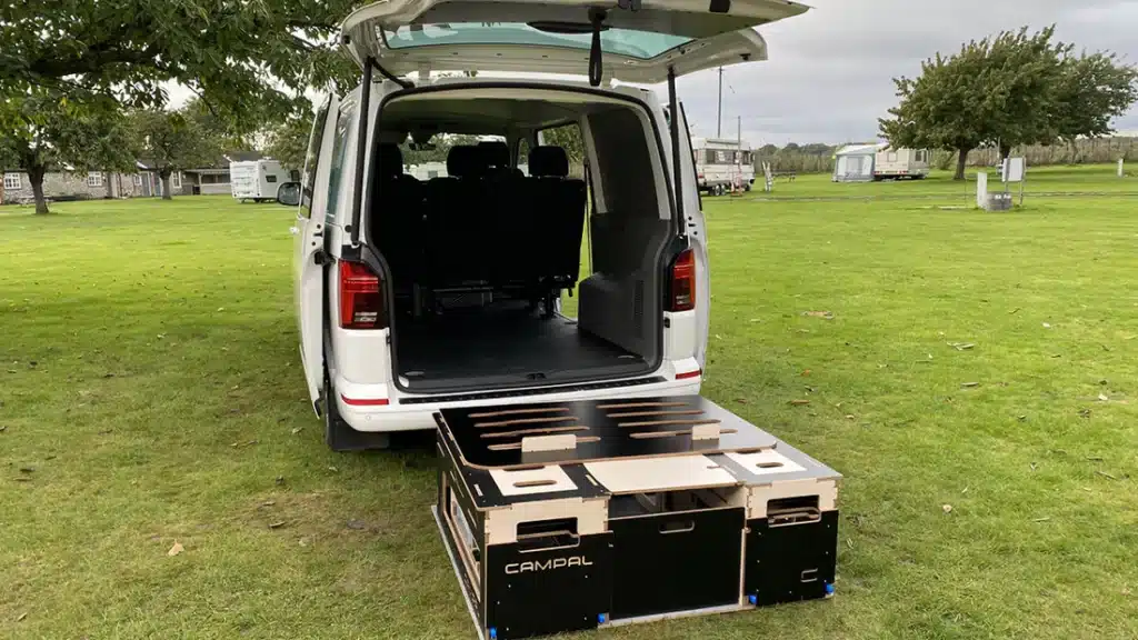 Standalone Campal camper box unit sitting on the grass outside an empty VW Transporter, demonstrating the system's fully removable design