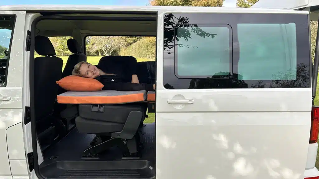 Side view of a VW Transporter T6.1 showing a woman lying on a Campal Elevate camper bed that extends over the folded rear Kombi seats.