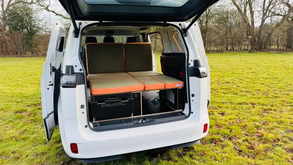 Campal removable camper conversion kit installed in a white VW ID Buzz boot, featuring orange cushions and birch plywood storage drawers with logo detail.