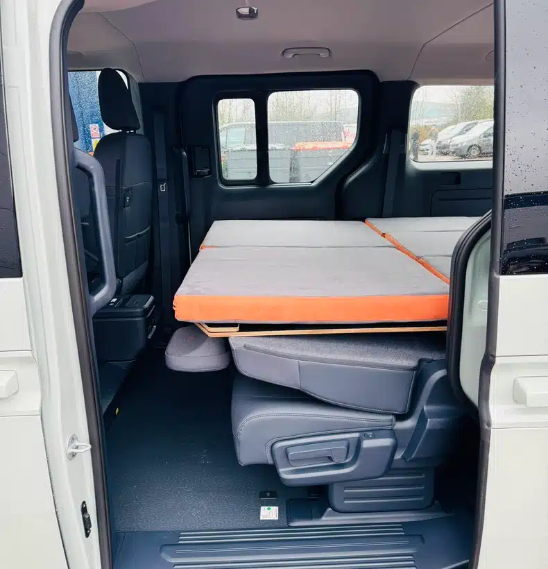 Side view through the sliding door of a Kia PV5 showing the standard 180cm removable flat camper bed deployed over folded passenger seats
