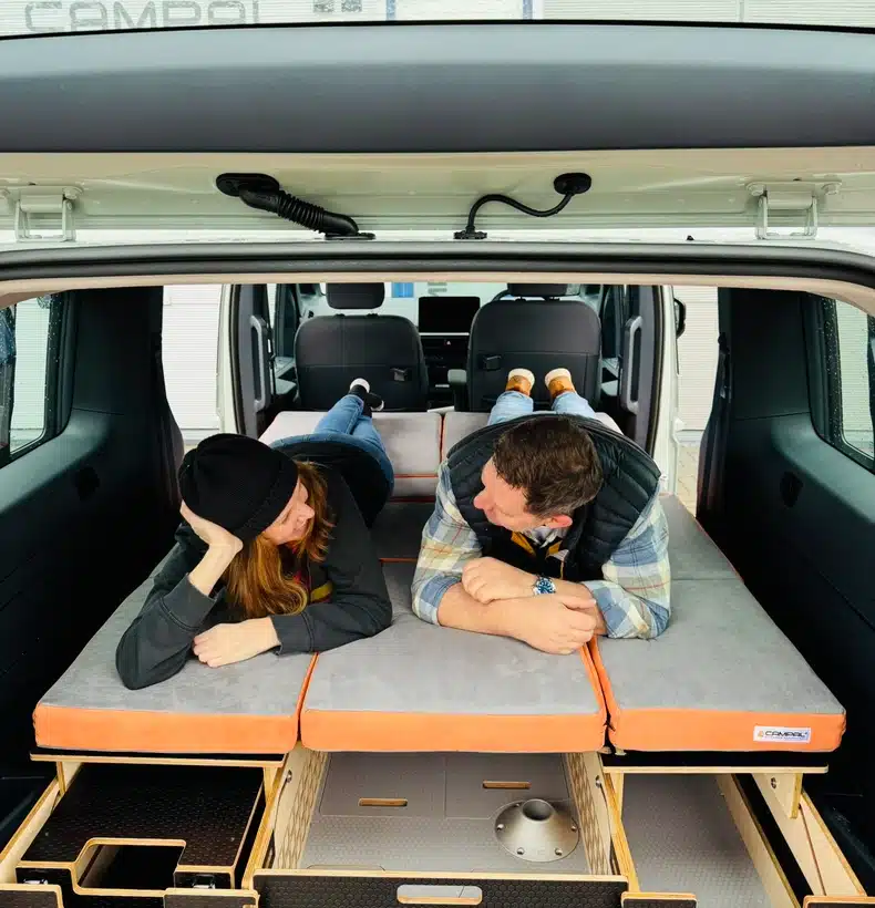 A couple relaxing comfortably on the fully flat, upholstered double bed inside their zero-emission Kia PV5 micro-camper