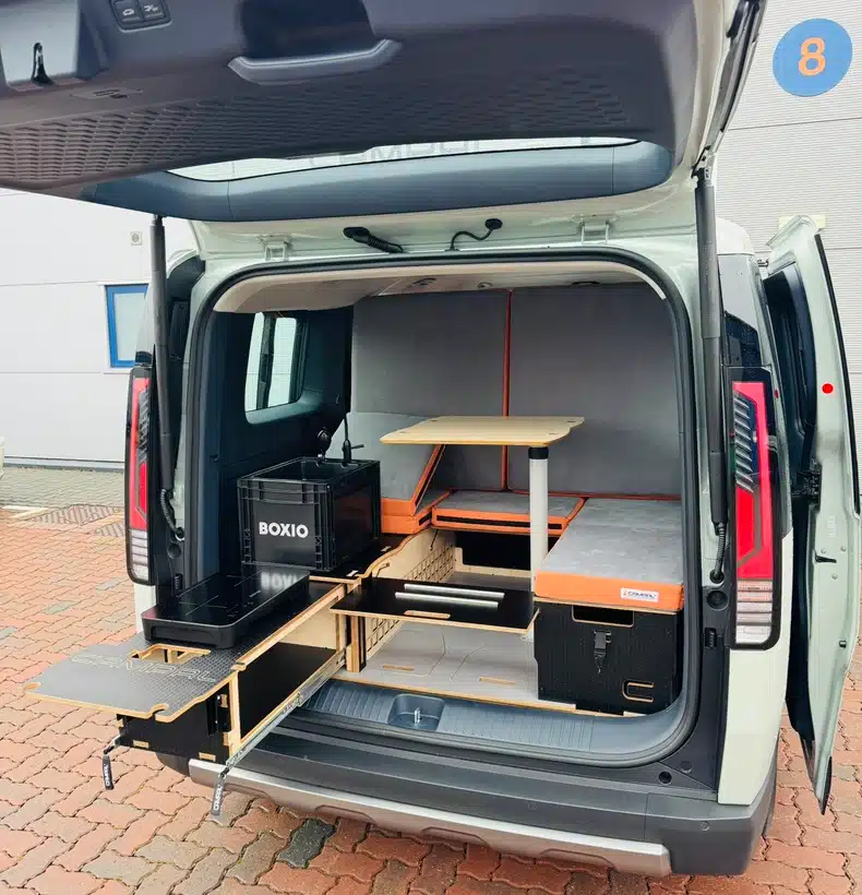 U-shaped seating and dining layout in the Kia PV5 camper featuring a central table and fully extended slide-out kitchen drawer with a portable sink