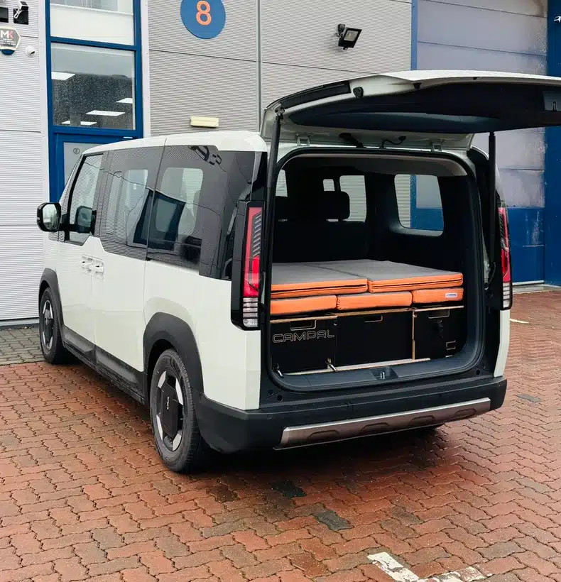 "Rear exterior view of a white Kia PV5 passenger vehicle with the boot open, revealing the neatly folded birch plywood Campal camper conversion kit inside