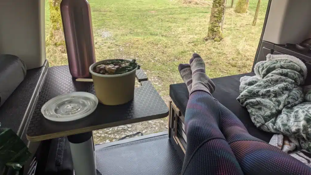 A person sitting on the edge of a Berlingo camper kit enjoying a cup of tea, with the tailgate providing a canopy over the pull-out kitchen.