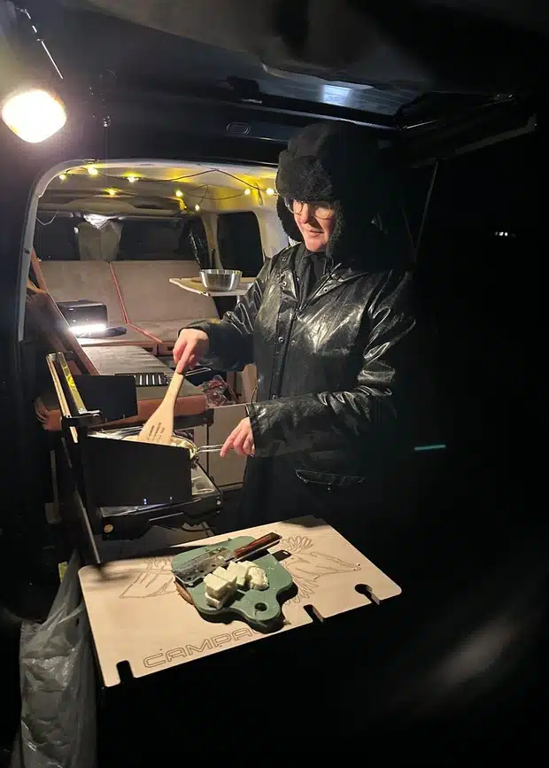 A person cooking dinner on a rear pull-out kitchen stove in a Citroën Berlingo car camper at night, featuring string lights for a cosy atmosphere.