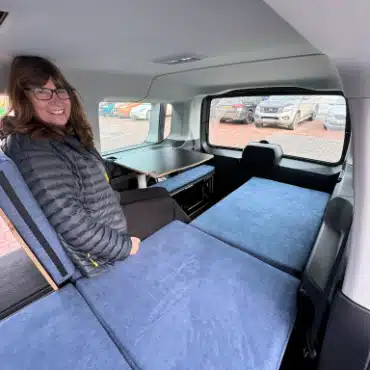 Woman sitting at indoor dining table inside VW Caddy micro camper demonstrating headroom and living space.