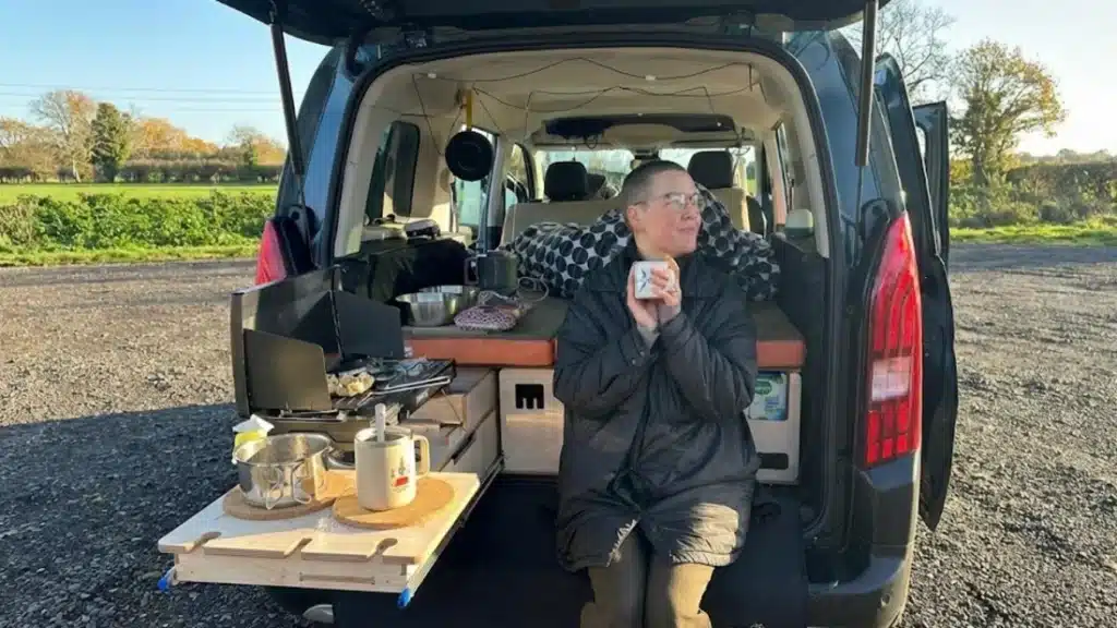 First-person view of someone relaxing on a black cushioned Berlingo camper bed with a meal on a swivel table, looking out at a green field.