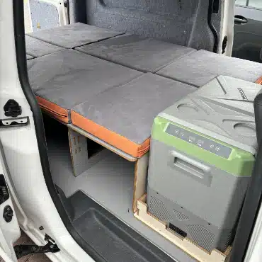 VW Caddy camper interior showing integrated storage solution for portable compressor fridge alongside sleeping platform.