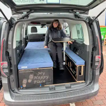 Woman sitting at indoor dining table in VW Caddy Maxi using Campal Split camper conversion kit with flexible seating layout and engraved storage.
