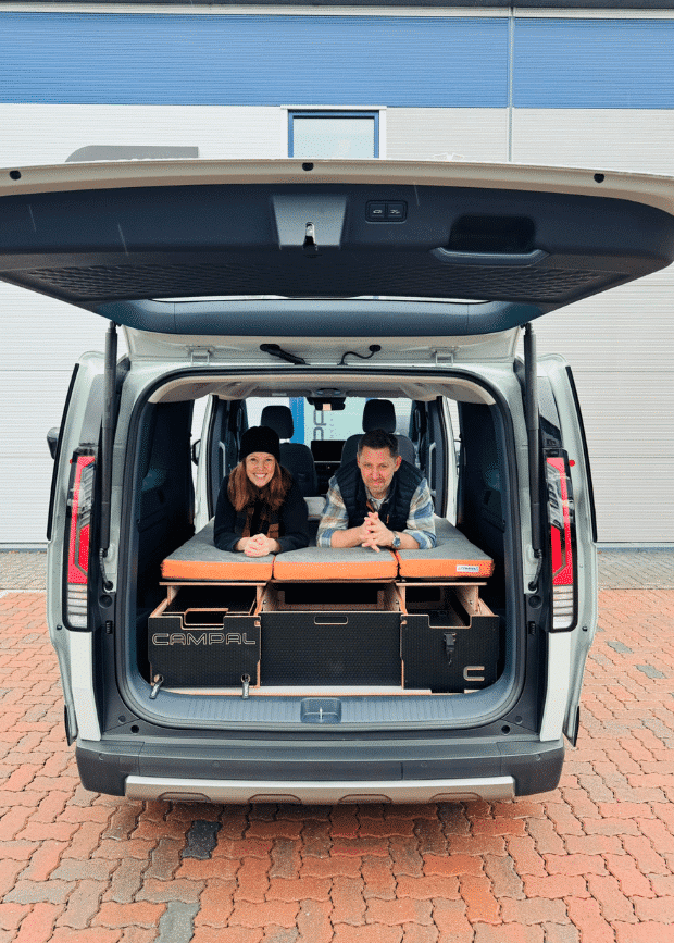 Lisa and Martin from Campal on the folded-out double bed of a Kia PV5 fitted with a removable Campal camper conversion kit.