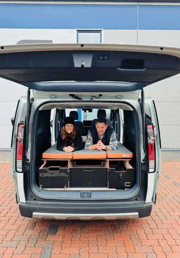 Lisa and Martin from Campal on the folded-out double bed of a Kia PV5 fitted with a removable Campal camper conversion kit.